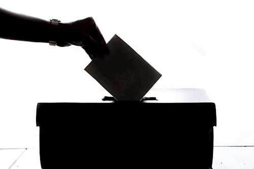 A hand wearing a watch placing a piece of paper in a ballot box. The hand paper, and box are in silhouette.