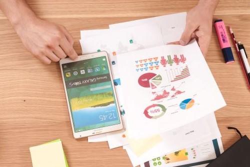 A pair of hands on a table. In between the hands are a tablet, papers with colorful graphs, and various office supplies